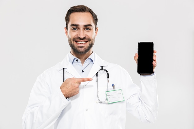 Portrait of cheery young medical doctor with stethoscope working in clinic and holding cellphone isolated over white wall