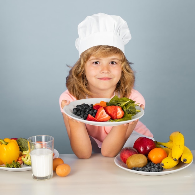 料理の帽子をかぶったシェフの子供の肖像画は、家で料理をしている果物と一緒にプレートを保持します子供の男の子はあちこちで食べ物を準備しています