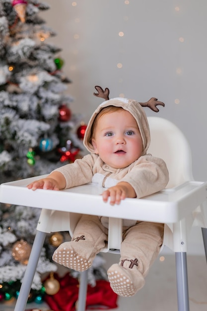 portrait of chubby baby in a jumpsuit with deer horns sits in a white high chair near Christmas tree