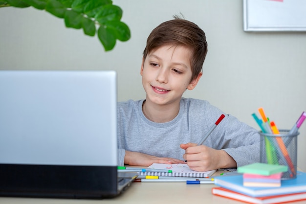 Portrait of clever schoolboy writing in notebook and using laptop while doing schoolwork at home sitting at desk.