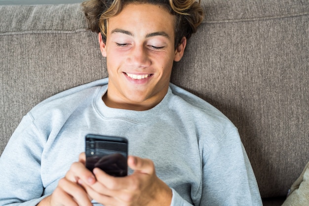 Portrait and close up of man teenager smiling and using his phone on the sofa of his home alone - social media and network - surfing on the net lifestyle and concept