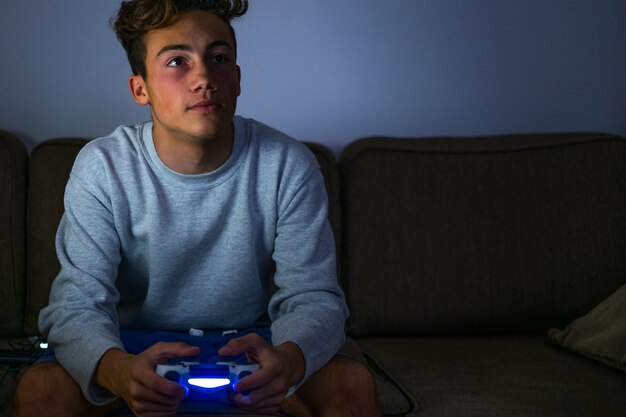 Портрет и крупный план подростка или миллениала, играющего в видеоигры с контроллером дома поздно ночью на диване - плохой нездоровый образ жизни большинства мальчиков