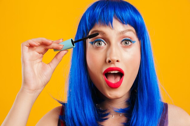 Portrait closeup of astonished woman wearing blue wig looking and applying mascara isolated over yellow wall