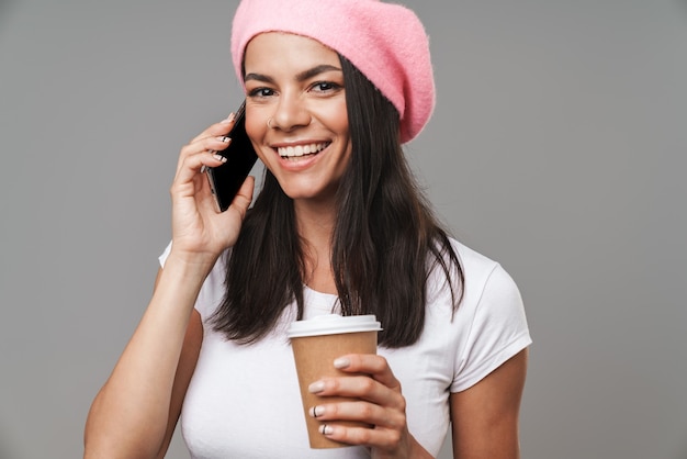 Primo piano del ritratto di bella donna graziosa in berretto che sorride e che parla sullo smartphone mentre beve caffè da asporto isolato sopra il muro grigio