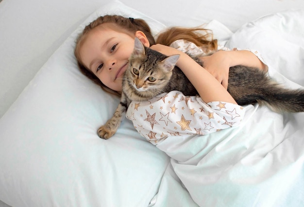 Portrait Closeup The child is resting on the bed with his kitten A little girl in pajamas on a sunny morning with tenderness and love hugs a gray cat and smiles