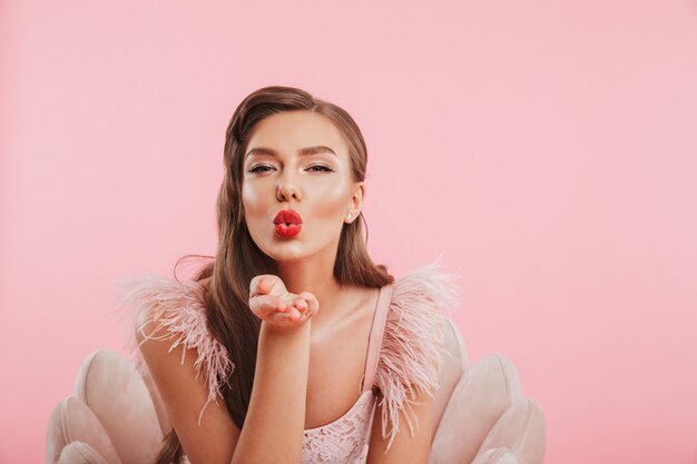 Portrait closeup of fancy seductive woman in dress sitting in armchair and giving air kiss to you, isolated over pink wall