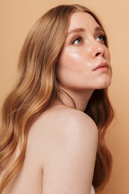 Portrait closeup of gorgeous young half-naked woman with long auburn hair looking aside at copyspace isolated on beige