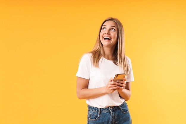 Primo piano del ritratto di una donna bionda felice che indossa una t-shirt casual che guarda verso l'alto il copyspace mentre utilizza lo smartphone isolato su un muro giallo