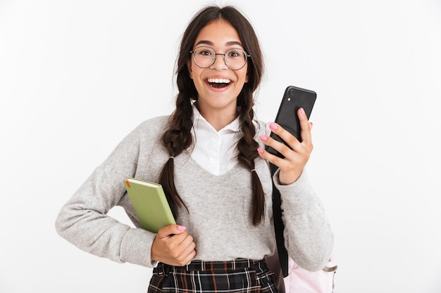 Portrait closeup of joyful teenage girl wearing eyeglasses laughing while holding studying books and smartphone isolated over white wall