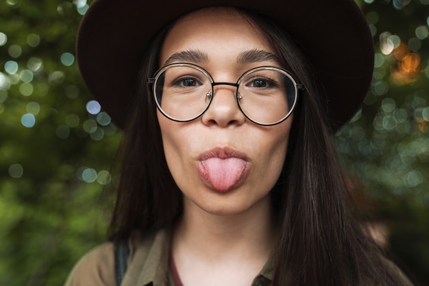 Portrait closeup of joyful woman wearing hat and eyeglasses showing her tongue while walking in evening park