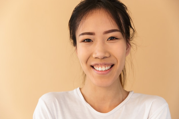 Portrait closeup of optimistic asian woman wearing basic t-shirt laughing and looking at front isolated over beige wall in studio