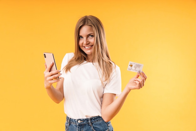 Foto primo piano del ritratto della donna bionda ottimista che indossa la maglietta casuale che sorride mentre tiene lo smartphone e la carta di credito isolati sopra la parete gialla