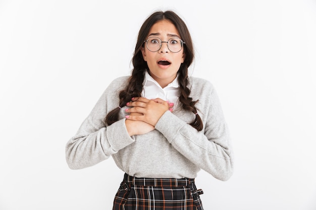 Portrait closeup of scared teenage girl wearing eyeglasses and school uniform screaming while touching her chest isolated over white wall