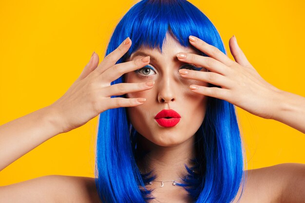 Portrait closeup of seductive beautiful woman wearing blue wig looking and covering her face isolated over yellow wall