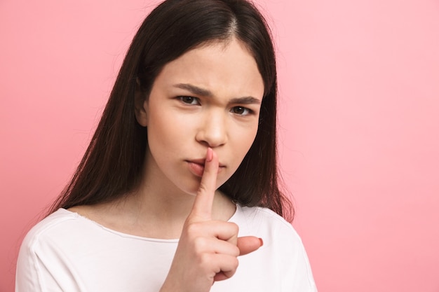Portrait closeup of serious girl with long dark hair holding index finger on lips and asking to be quiet isolated over pink wall