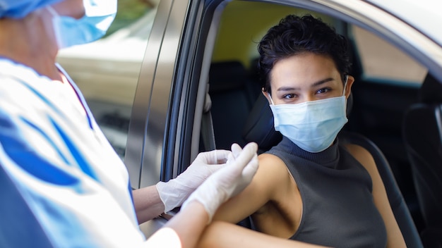 Photo portrait closeup shot of female wearing face mask sitting in car receiving coronavirus vaccine from doctor wears hospital uniform using syringe and needle in drive through vaccination queue.