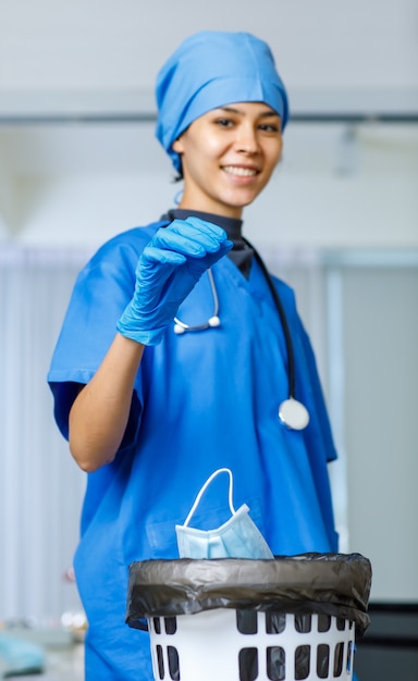 Portrait closeup shot of used surgical face mask dumped drop\
from happy pretty female freedom doctor hand in blue hospital suit\
hat with stethoscope smiling look at camera while covid pandemic\
end.