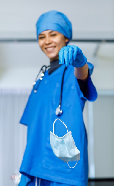 사용된 수술용 얼굴 마스크의 초상화 클로즈업 샷은 코비드 전염병이 끝나는 동안 청진기 미소가 카메라를 바라보는 파란색 병원 양복 모자에 행복한 예쁜 여성 자유 의사의 손에서 한 방울을 버렸다.