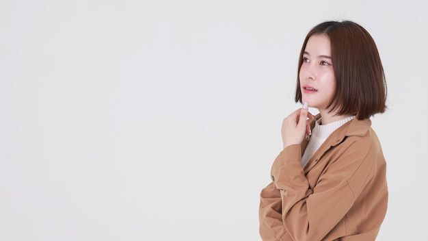 Portrait closeup studio cutout shot of Asian young beautiful short hair female model in brown coat jacket standing smiling look at camera pointing finger up on blank copy space on white background.