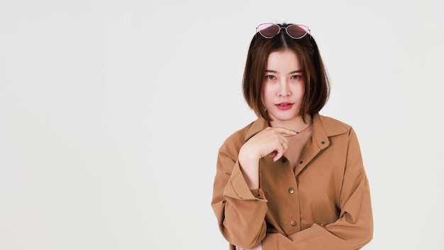 Portrait closeup studio cutout shot of Asian young beautiful short hair female model in brown coat jacket standing smiling look at camera pointing finger up on blank copy space on white background.