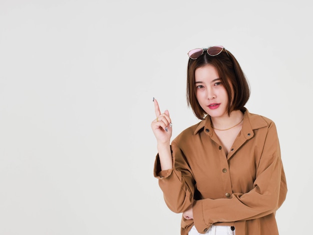 Portrait closeup studio cutout shot of Asian young beautiful short hair female model in brown coat jacket standing smiling look at camera pointing finger up on blank copy space on white background.