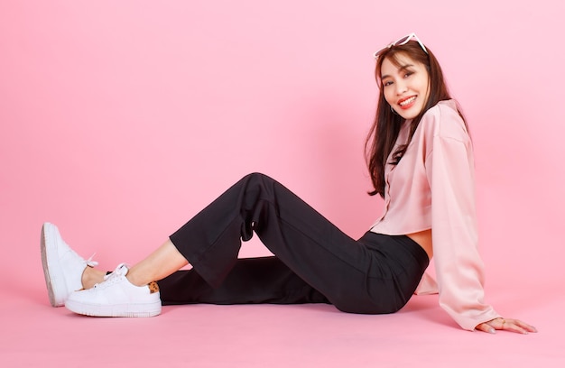 Portrait closeup studio shot of Asian young beautiful hipster female teenager model in casual long sleeve shirt street wears and sunglasses sitting posing smiling look at camera on pink background.