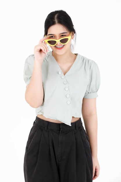 Portrait closeup studio shot of Asian young cool stylish teen female model with ponytail hairstyle in casual crop top outfit holding hand touching fashionable sunglasses wink eye on white background.