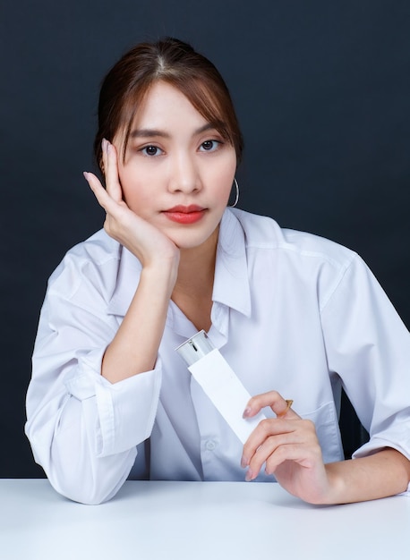 Portrait closeup studio shot of Asian young glamour trendy fashionable female model wearing makeup in casual white shirt sitting smiling look at camera holding face cream bottle on black background