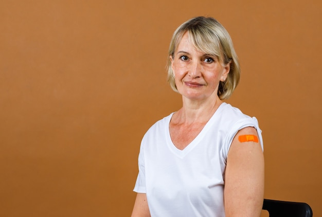 Portrait Closeup studio shot of Caucasian senior blonde female patient stand smiling look at camera show orange plaster bandage on her shoulder after receive Coronavirus Covid-19 vaccine injection.