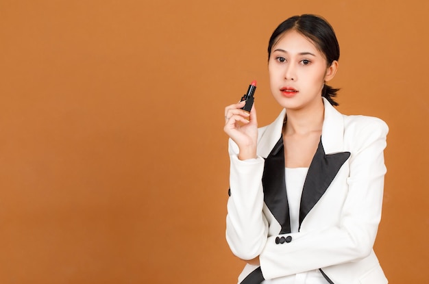 Portrait closeup studio shot millennial Asian black short ponytail hair female model wears makeup in black and white fashion casual blazer crossed arm holding red lipstick in hand on brown background.