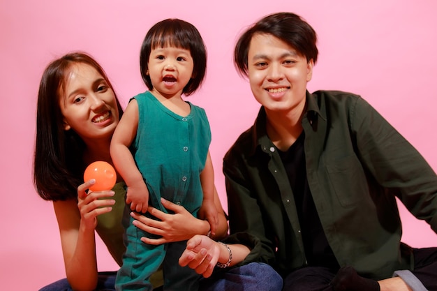 Portrait closeup studio shot of young happy Asian family father mother and little cute baby girl daughter sitting on floor together smiling look at camera holding colorful balloons on pink background.