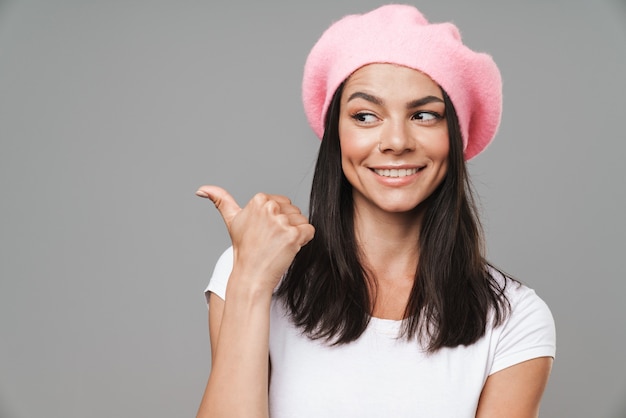 Portrait closeup of surprised pretty woman in basic t-shirt and beret smiling while pointing finger aside at copyspace isolated over gray wall