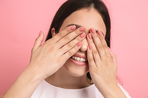 Portrait closeup of sweet joyful girl with long dark hair covering her eyes and peeking though hands isolated over pink wall