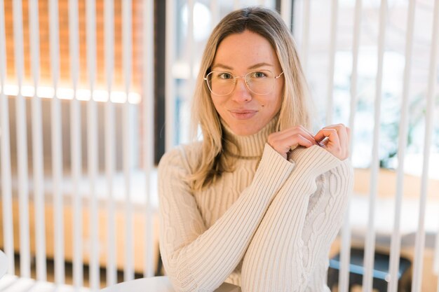 Portrait of comforting happy cozy woman in glasses in knitted winter white warm sweater in cafe