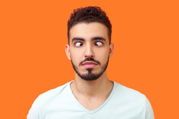 Portrait of comic positive brunette man with beard in casual white tshirt looking crosseyed having fun with silly face expression playing fool indoor studio shot isolated on orange background