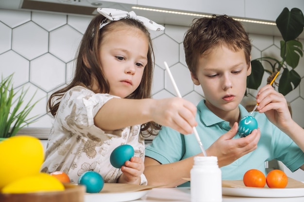 Ritratto di concentrato gioioso due bambini ragazzino e ragazza pennello pittura multi colorazione uova di galline pasquali su tavola di piatto di carta fare modelli di design d'arte bianca in cucina giorno di luce primo piano