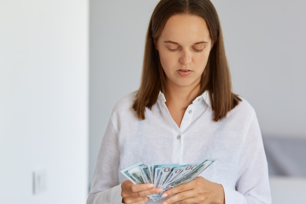 Portrait of concentrated woman counting money at home, dark haired female standing with dollar banknotes in hands, wearing white shirt, posing in light room.