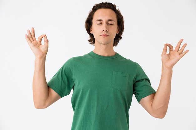 Photo portrait of concentrated young man wearing basic t-shirt keeping fingers together in meditation isolated over white wall