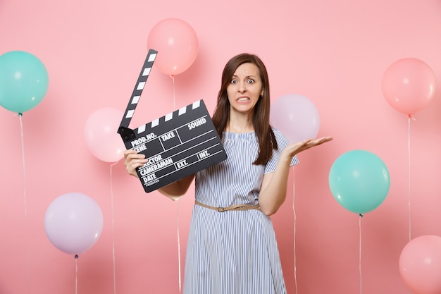 Portrait of concerned dissatisfied woman wearing blue dress spreading hands holding classic black film making clapperboard on pink background with colorful air baloons. Birthday holiday party concept.