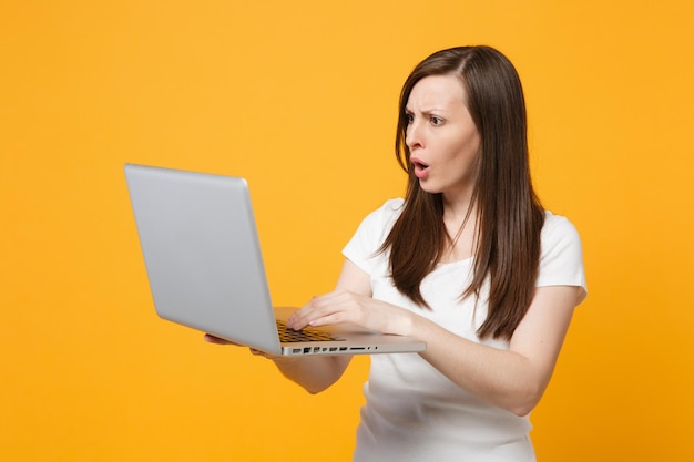 Portrait of concerned puzzled young woman in white casual clothes using laptop pc computer isolated on bright yellow orange wall background in studio. People lifestyle concept. Mock up copy space.