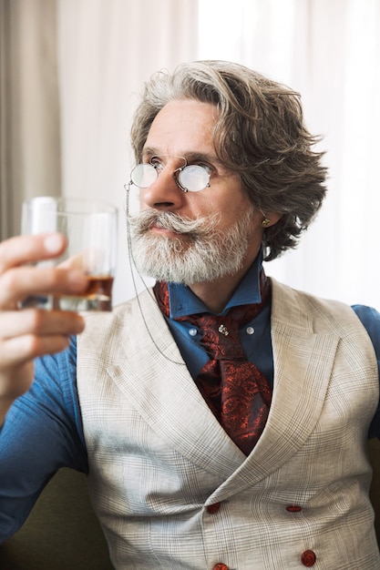 Portrait of confident adult businessman wearing stylish suit drinking alcohol while sitting on armchair in hotel apartment