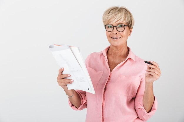 Portrait of confident adult woman wearing eyeglasses holding studying book and pen isolated over white wall in studio