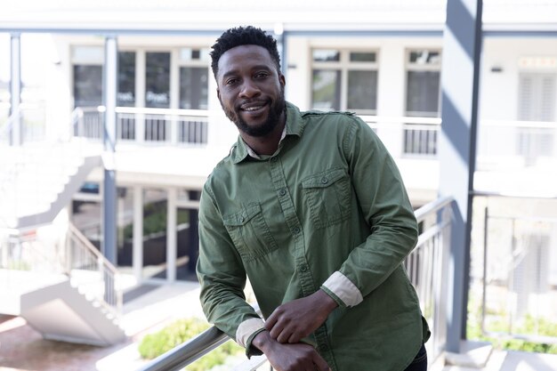 Photo portrait of confident african american businessman in a modern workplace standing on a balcony in sun, leaning on handrail smiling. social distancing in workplace during coronavirus covid 19 pandemic.