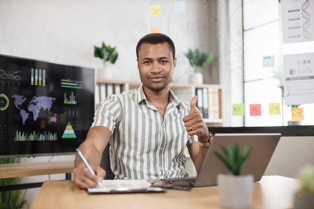 Portrait of confident african businessman in stylish formal wear writing notes using laptop