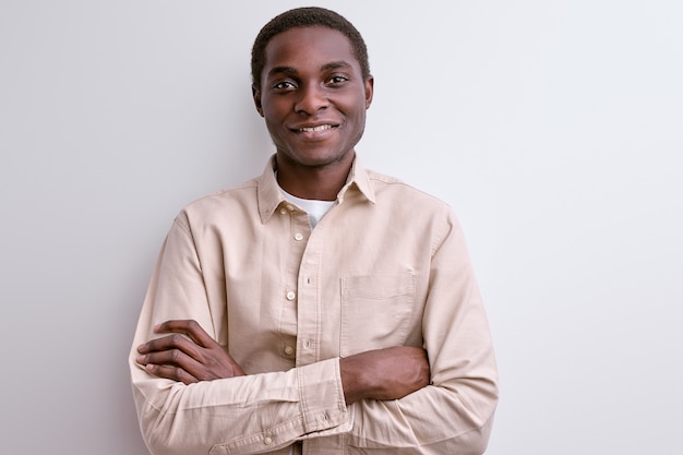 Photo portrait of confident african guy standing with arms folded and smiling at camera, wearing light-colored clothing isolated on white studio wall