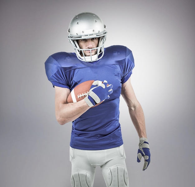 Portrait of confident American football player holding ball against grey background