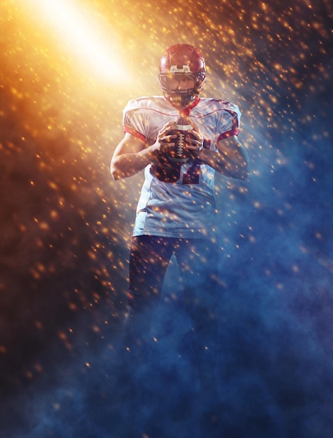 portrait of confident American football player holding ball while standing on the big field with particles effects and lights