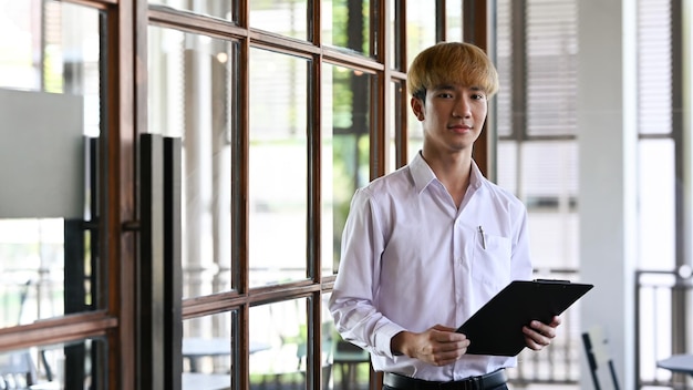 Портрет уверенного азиатского работника-мужчины, держащего буфер обмена и стоящего в современном офисе
