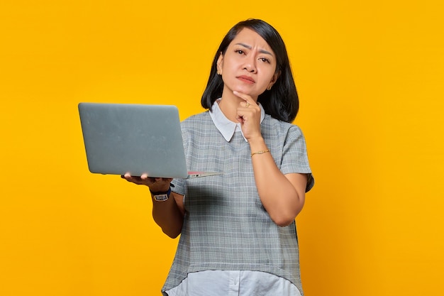 Portrait of confident Asian woman holding laptop and looking at camera over yellow background