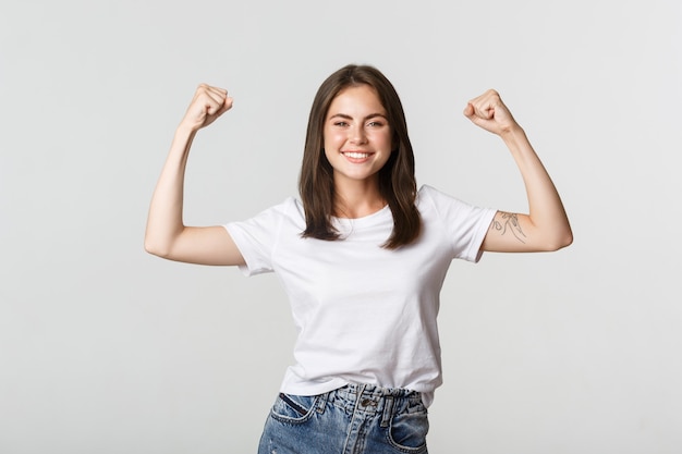 Portrait of confident attractive girl flex biceps to show strengths, smiling sassy on white.